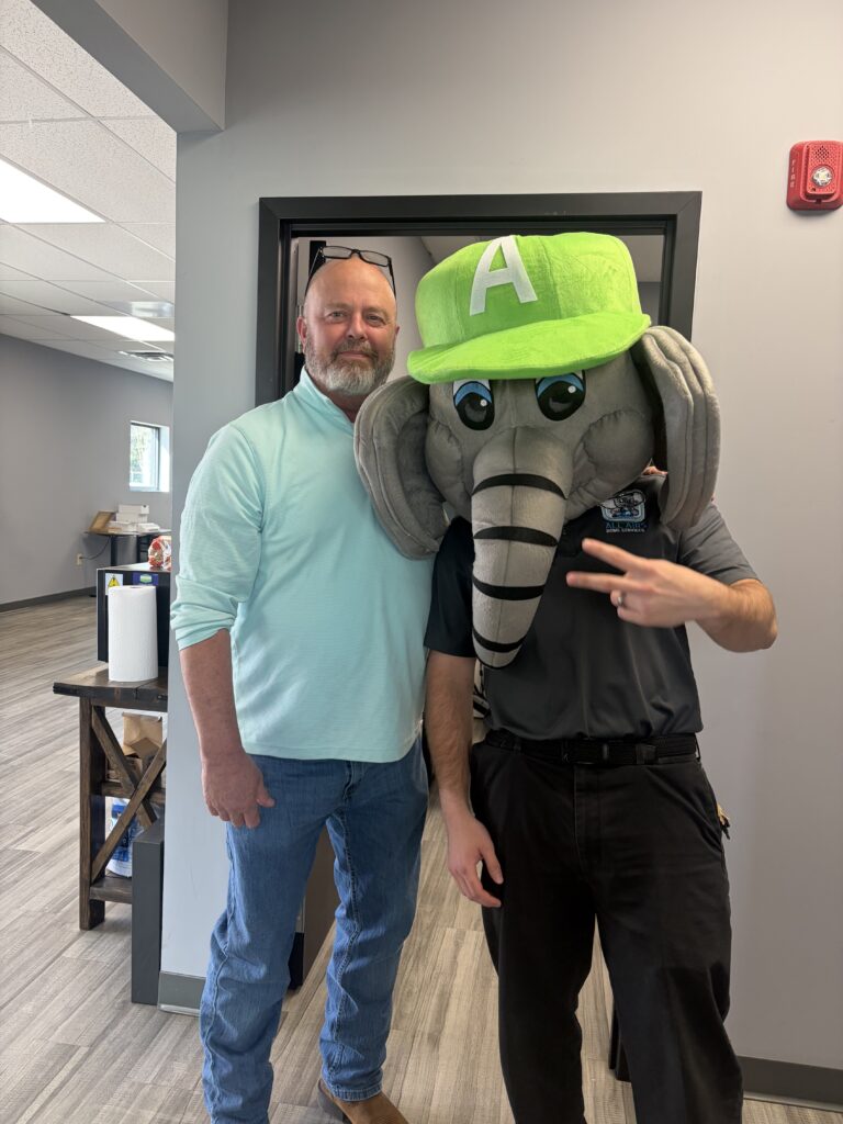 Two men pose indoors; one in a teal shirt, the other wearing an elephant mascot head with a bright green hat marked with the letter A.