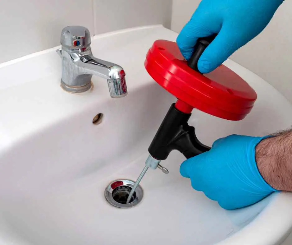 Gloved hands operate a red manual drain auger to unclog a bathroom sink drain.